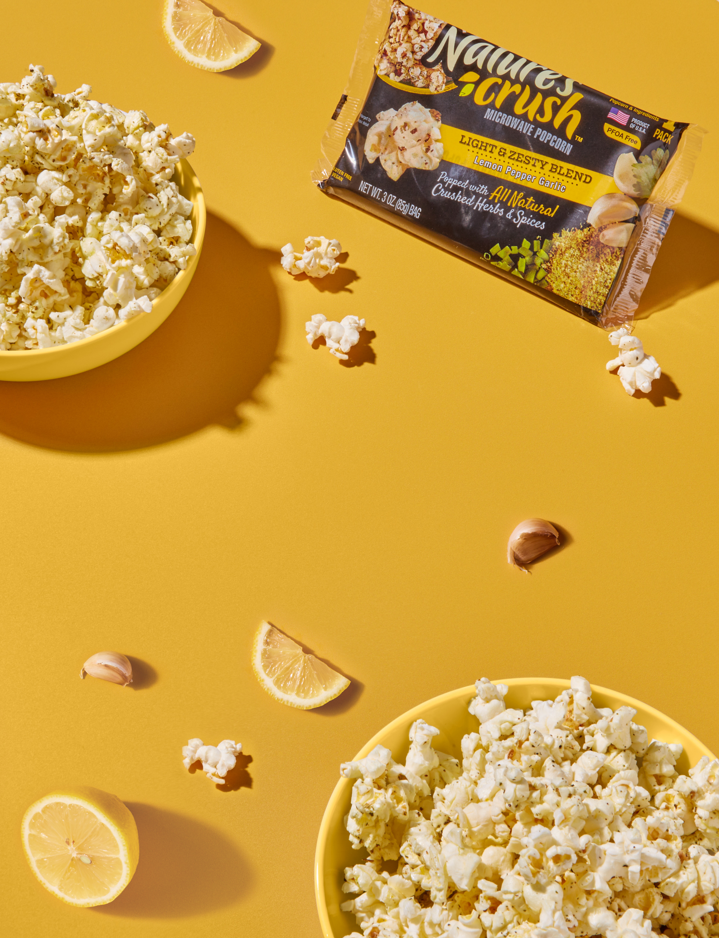 Bag and bowls of Light & Zesty popcorn surrounded by scattered popcorn, lemons and garlic on yellow surface.