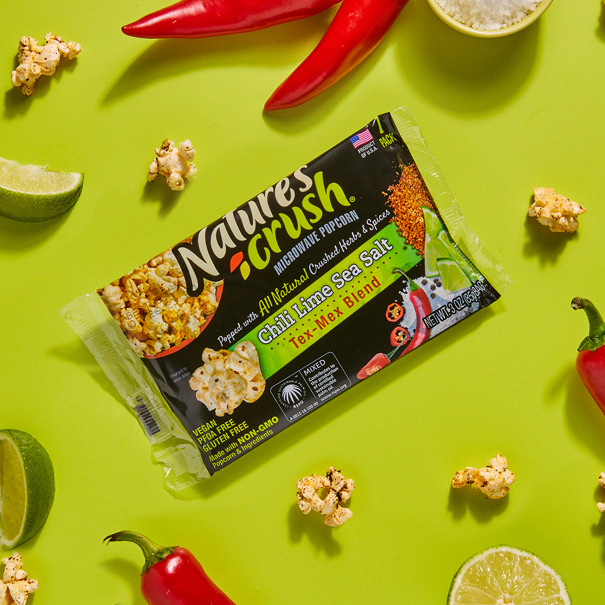 Bag of Tex-Mex popcorn surrounded by scattered popcorn, peppers and limes on green background.